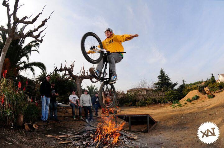 A rider in a yellow XAI hoodie jumping his bike over a bonfire while friends watch