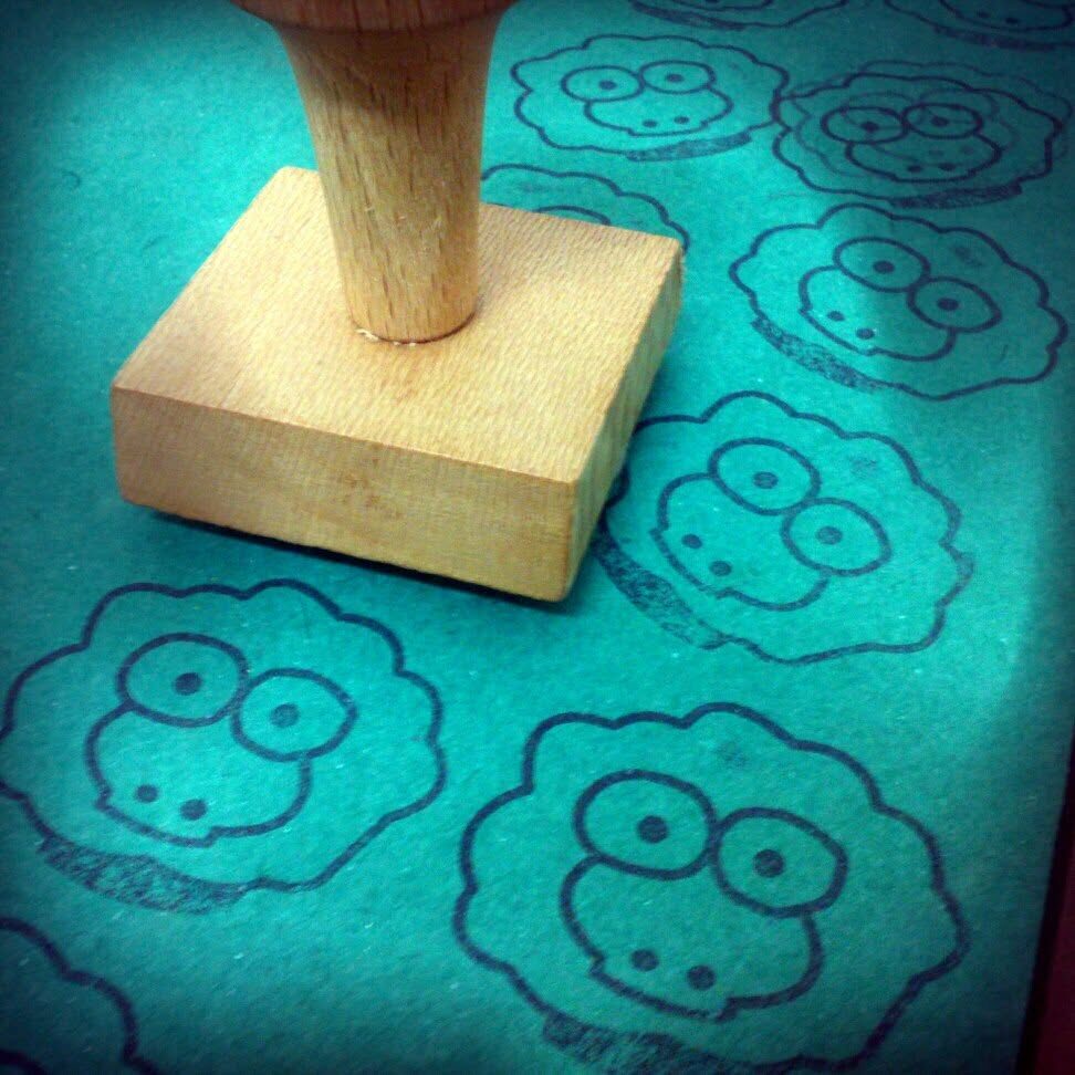 A wooden rubber stamp resting on teal paper printed with rows of stamped sheep